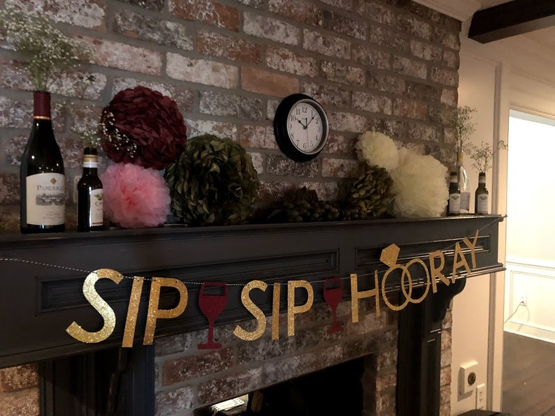 Puede incluir: Una pancarta de purpurina dorada con el texto "Sip Sip Hooray" y un anillo dorado con un diamante. La pancarta est&aacute; colgada sobre una chimenea con un marco de ladrillo. Tambi&eacute;n hay algunas bolas de papel decorativas en el fondo.