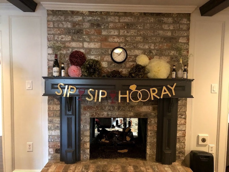 Puede incluir: Una chimenea de ladrillo decorada con una pancarta dorada que dice "Sip Sip Hooray" con dos anillos dorados. La chimenea est&aacute; decorada con botellas de vino, pompones de papel de seda rosa y amarillo y vegetaci&oacute;n.
