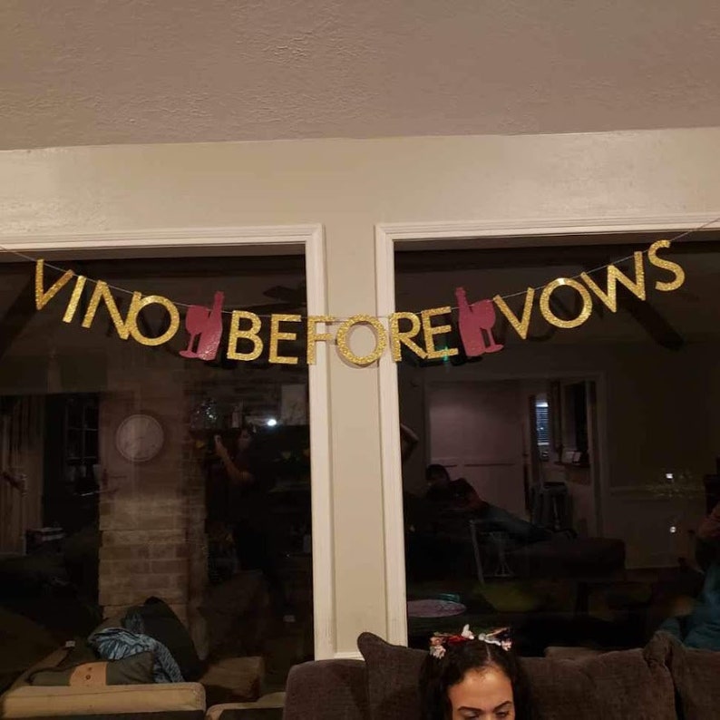 Puede incluir: Pancarta de purpurina dorada con el texto "VINO BEFORE VOWS" y siluetas de copas de vino rosas.