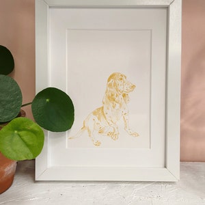 May include: A framed illustration of a golden basset hound dog sitting with its head turned to the side. The dog is outlined in gold and has a white background.