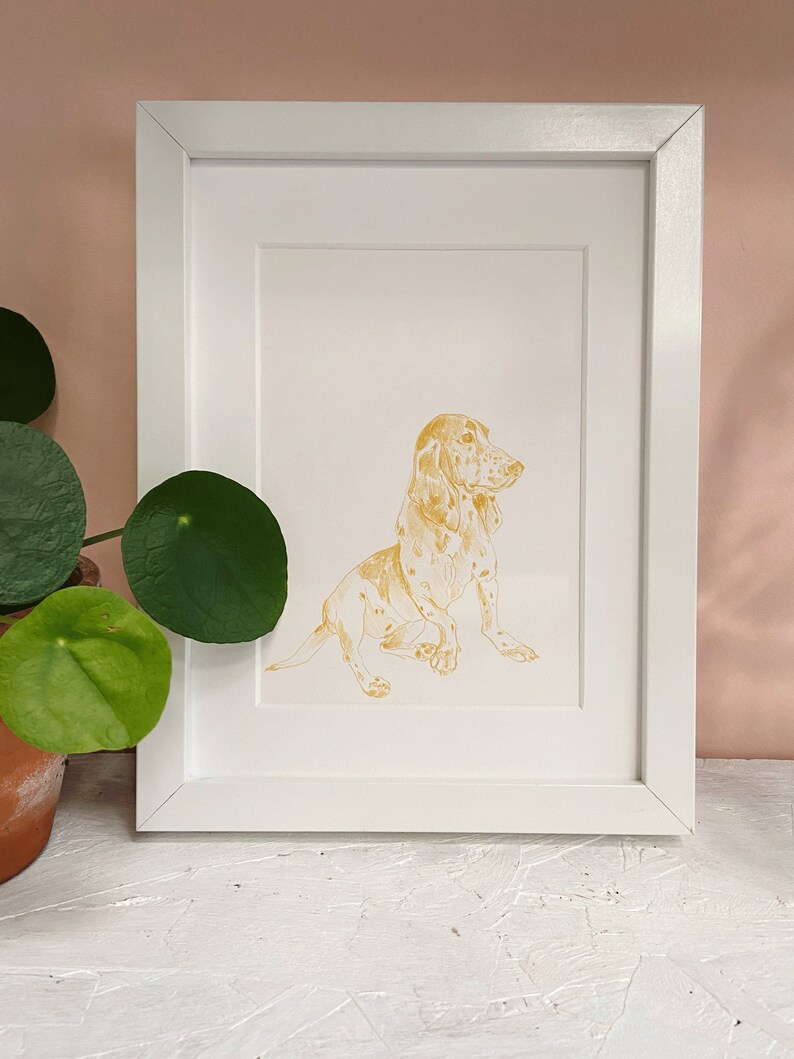 May include: A framed illustration of a golden basset hound dog sitting with its head turned to the side. The dog is outlined in gold and has a white background.