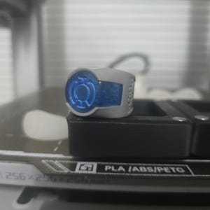Pode incluir: Um anel cinzento com um emblema circular azul proeminente com um símbolo central. O anel tem um acabamento mate liso e está posicionado sobre uma superfície preta com o texto PLA /ABS/PETG e 256x256mm visível na base.