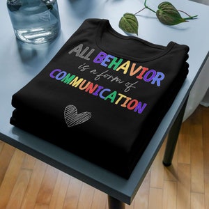 Könnte beinhalten: Schwarzes T-Shirt mit dem Text "ALL BEHAVIOR is a form of COMMUNICATION" in Regenbogenfarben. Ein kleines graues Herz befindet sich unter dem Text.