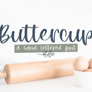 May include: A close-up of the word "Buttercup" written in a blue, hand-lettered font. The text is on a white background with a green and white line below it that says "A hand lettered font". There are three eggs in a white bowl and a wooden rolling pin on a white surface.