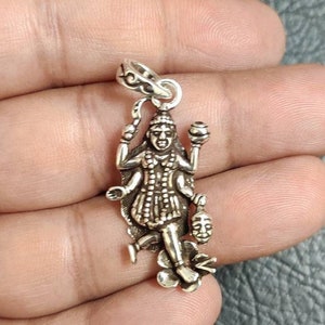 Può includere: Un ciondolo d'argento raffigurante una divinità con più braccia e dettagli intricati. Il ciondolo include elementi floreali e un piccolo ciondolo. La figura tiene in mano vari oggetti e il ciondolo ha un anello per una catena.