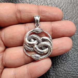 以下が含まれることがあります： 2匹の蛇が絡み合って円形になっているシルバー製のペンダント。蛇は鱗模様で、リアルな目つきをしている。