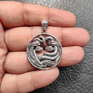 May include: A silver pendant featuring two intertwined snakes in a circular design. The snakes are detailed with scales and have their heads facing each other.