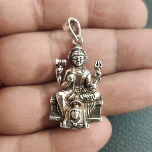 Könnte beinhalten: Silberner Anhänger mit einer detaillierten Darstellung einer hinduistischen Gottheit, wahrscheinlich Lakshmi, die auf einem Thron mit einem Löwen zu ihren Füßen sitzt. Sie hält eine Lotusblume und andere Symbole in ihren Händen.