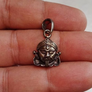 Può includere: Ciondolo in argento con una rappresentazione dettagliata di una divinità con un'espressione feroce, che indossa una corona e una lunga lingua. Il ciondolo ha un anello nella parte superiore per attaccarlo a una catena.