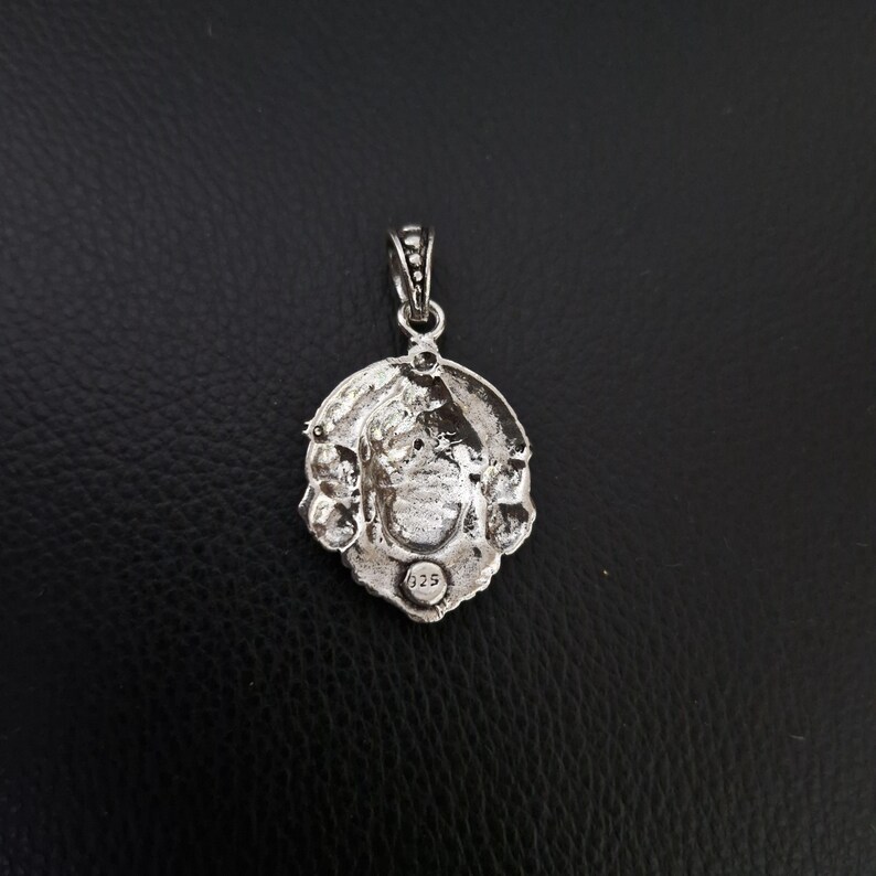 May include: Silver pendant featuring an intricate design of a face surrounded by floral motifs. The reverse of the pendant is stamped with the number "925".
