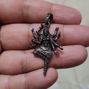 Può includere: Un ciondolo d'argento raffigurante una divinità con molte braccia in posa danzante. Il ciondolo presenta dettagli intricati, con ogni braccio e tratto del viso chiaramente definito. L'ansa è un semplice anello per attaccare una catena.