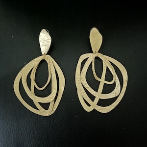 Puede incluir: Un par de pendientes colgantes de color dorado con un diseño abstracto en capas. Los pendientes tienen una textura martillada y están hechos de metal.