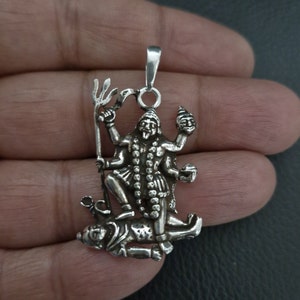 Può includere: Ciondolo in argento con una rappresentazione dettagliata della divinità indù Kali, in piedi su un demone. Kali è raffigurata con quattro braccia, che tengono un tridente, un teschio e una ciotola. Il ciondolo è finemente lavorato e presenta un design indiano tradizionale.