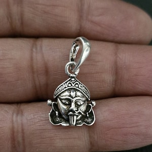 Può includere: Ciondolo in argento con una rappresentazione dettagliata del volto di una divinità con la lingua sporgente. Il ciondolo ha un anello nella parte superiore per attaccarlo a una catena.