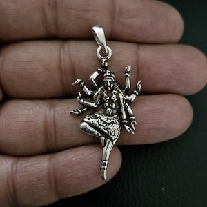 Può includere: Un ciondolo d'argento raffigurante una divinità multibrachiale in una posa dinamica. Il ciondolo ha un anello nella parte superiore per una catena. La divinità è adornata con dettagli intricati, tra cui gioielli e abbigliamento.