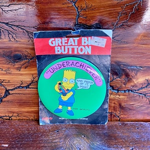 May include: A green button with a cartoon image of Bart Simpson from The Simpsons. Bart is holding a slingshot and the text on the button reads "Underachiever and proud of it, man!"  Matt Groening is credited as the creator.
