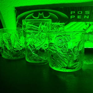 May include: Three clear glass mugs and a tumbler with embossed images of Batman characters. The mugs feature the Joker, Batman, and other characters. The background includes a Batman logo and the words "Poster Pens".