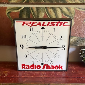 May include: A square wall clock with a white face, black numerals, and black hands. The top features the red text REALISTIC, and the bottom features the red text Radio Shack. The clock has a dark frame and includes the Lux brand logo.