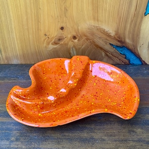 May include: An orange ceramic dish with an irregular, kidney-like shape and a glossy finish. The surface is decorated with a speckled pattern of small red and yellow dots. The dish features a raised central divider that creates two distinct sections.