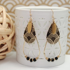 May include: Gold teardrop earrings with black beads and a geometric pattern on the teardrop. The earrings are hanging from a white ceramic cup.