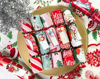 May include: A collection of cylindrical Christmas crackers arranged on a gold-rimmed plate. The crackers feature festive patterns including red and white candy stripes, snowmen, Santa Claus, and text reading Merry Christmas and Let It Snow.