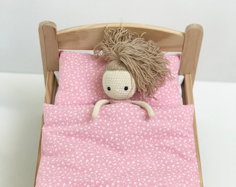May include: A wooden doll's bed with a pink floral patterned bedding set. A crocheted doll with brown hair is lying in the bed.