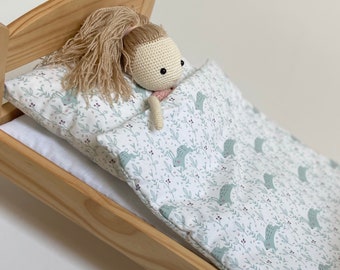 May include: A wooden doll's bed with a white and green patterned bedding set. A crocheted doll with blonde hair is lying in the bed.