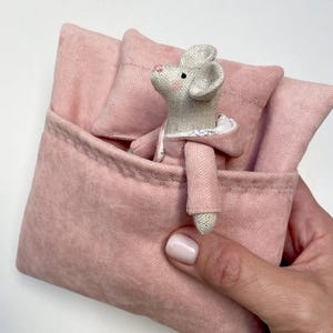 Puede incluir: Un pequeño ratón de peluche gris que lleva una bata rosa con capucha está metido en una cama de lona rosa con una almohada.