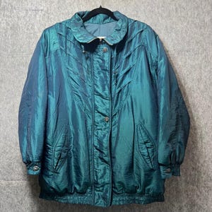 May include: Teal-colored, iridescent jacket with a high collar and button-up front. The jacket has a quilted design on the front and sleeves, with two front pockets. The jacket is hanging on a black hanger against a gray background.