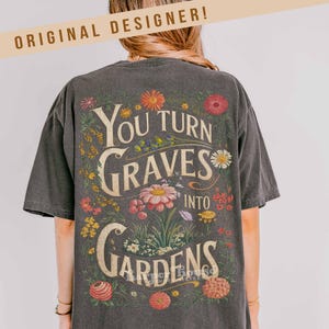 May include: Dark gray t-shirt with the text "You Turn Graves Into Gardens" surrounded by colorful flowers. The shirt has a vintage, worn look and the words "Original Designer" are at the top.
