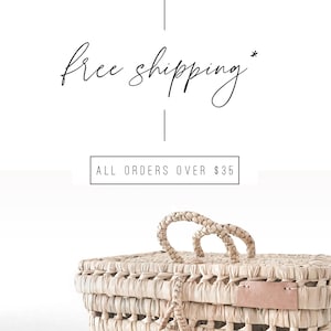 Op de afbeelding: Een geweven mand met een lichtbruine kleur en twee handvatten. De mand heeft een roze leren label aan de zijkant. De tekst "free shipping* ALL ORDERS OVER $35" is zichtbaar in de afbeelding.