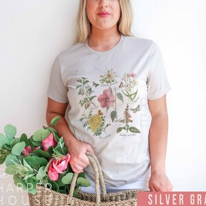 以下が含まれることがあります： 花と蝶の植物画が描かれたシルバーグレーのTシャツ。様々な花の種の詳細なイラストが、それぞれのラテン名とともにデザインされています。このシャツはニュートラルカラーで、普段使いに適しています。
