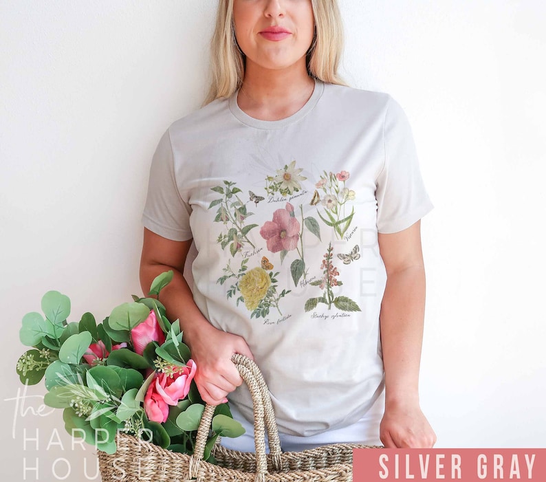 以下が含まれることがあります： 花と蝶の植物画が描かれたシルバーグレーのTシャツ。様々な花の種の詳細なイラストが、それぞれのラテン名とともにデザインされています。このシャツはニュートラルカラーで、普段使いに適しています。