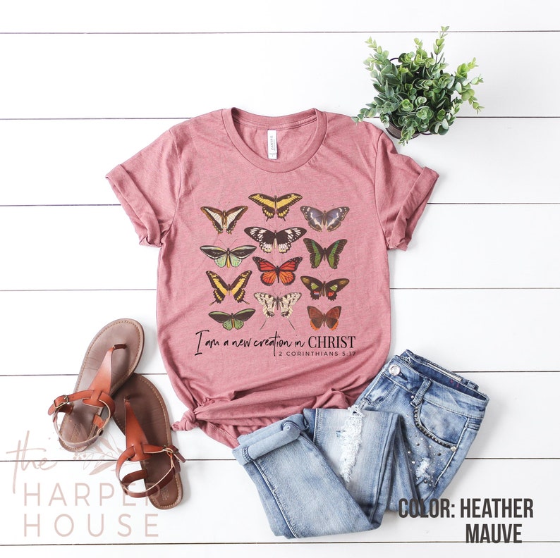 Puede incluir: Una camiseta de color malva brezo con un gr&aacute;fico de mariposas de colores y el texto "Soy una nueva creaci&oacute;n en CRISTO 2 Corintios 5:17".