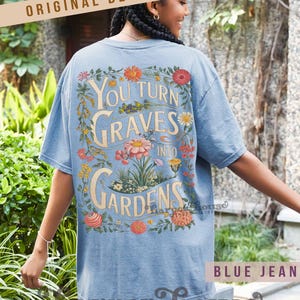 May include: A light blue t-shirt with the text "You Turn Graves Into Gardens" in a floral design. The shirt has short sleeves and a relaxed fit. The words "Original Designer" are printed at the top.