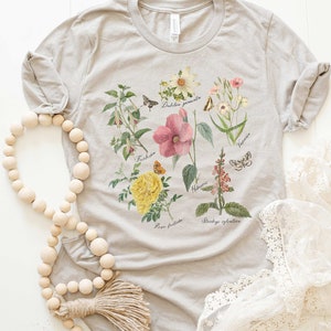 以下が含まれることがあります： 様々な花と蝶の植物画が描かれたシルバーグレーのTシャツ。ピンクのハイビスカス、黄色のバラ、その他の植物要素と学名が含まれています。Tシャツは丸いネックラインと半袖です。