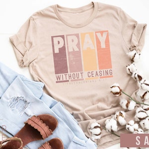 Op de afbeelding: Een beige T-shirt met de woorden "Pray Without Ceasing" en een bijbelvers in een vintage lettertype. Het shirt ligt plat op een wit oppervlak met een blauwe jeans, bruine sandalen en een tak katoen.