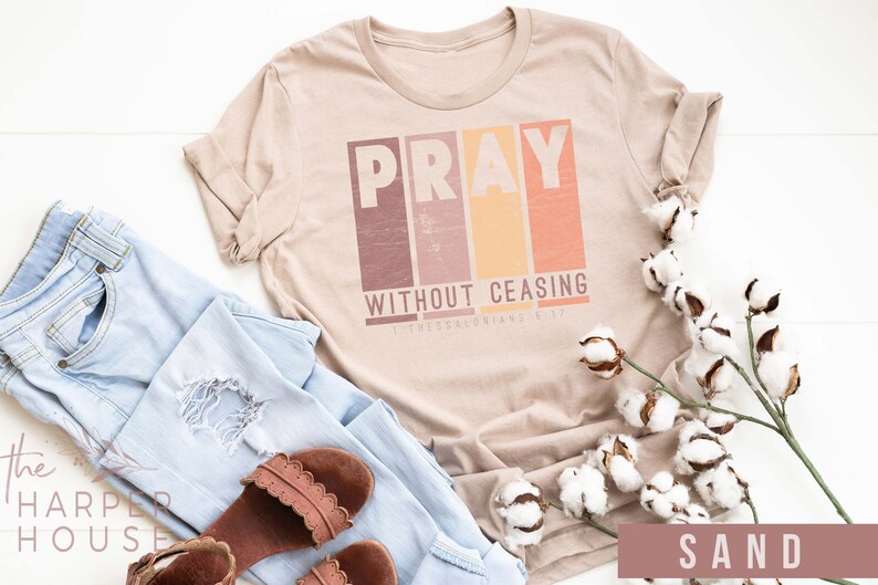 Op de afbeelding: Een beige T-shirt met de woorden "Pray Without Ceasing" en een bijbelvers in een vintage lettertype. Het shirt ligt plat op een wit oppervlak met een blauwe jeans, bruine sandalen en een tak katoen.