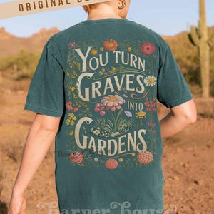 May include: Dark teal t-shirt with a floral design and the text "You Turn Graves Into Gardens." The design features various flowers and foliage. The words "Original Designer" are printed at the top. The shirt is worn by a person.