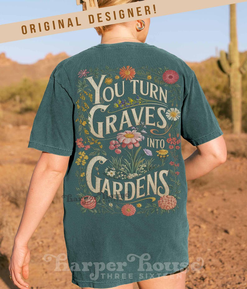 May include: Dark teal t-shirt with a floral design and the text "You Turn Graves Into Gardens." The design features various flowers and foliage. The words "Original Designer" are printed at the top. The shirt is worn by a person.
