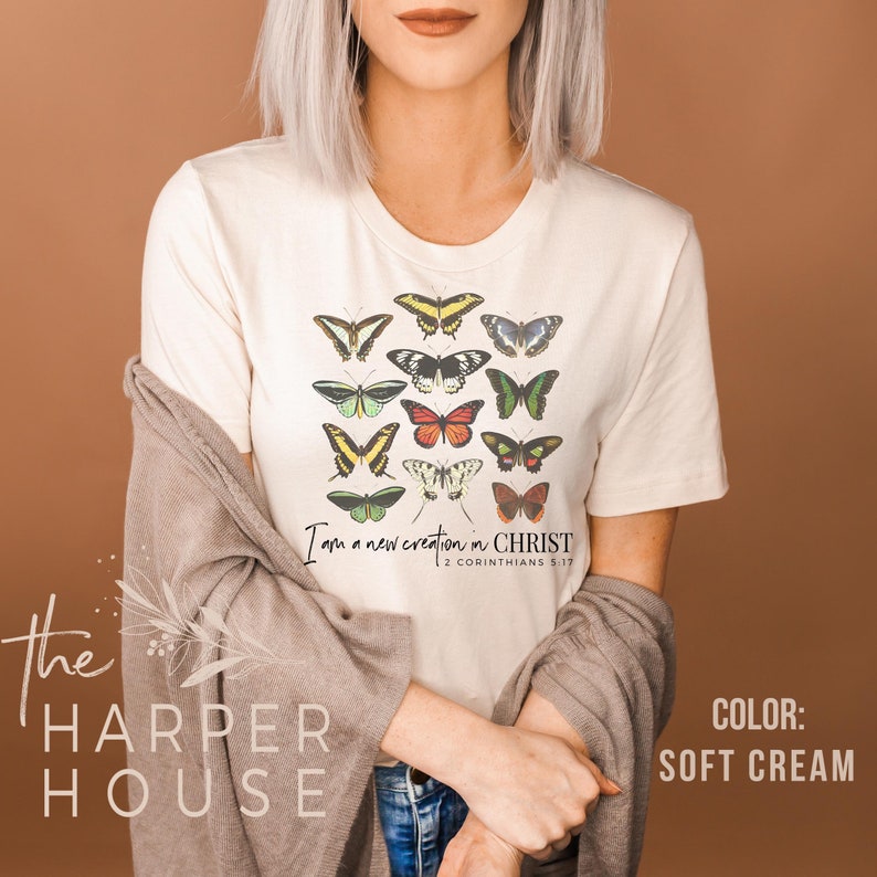 Puede incluir: Una camiseta blanca con un dise&ntilde;o de mariposas de colores y el texto "Soy una nueva creaci&oacute;n en CRISTO 2 CORINTIOS 5:17".