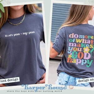 May include: A dark gray t-shirt with the text "God's plan > my plan" on the front and the text "do more of what makes you GOD happy" on the back. The text on the back is in a colorful, retro font.