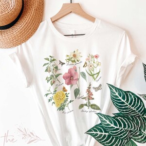 以下が含まれることがあります： 様々な花と蝶の植物画が描かれた白いTシャツ。学名が記載されています。Tシャツはクルーネックで半袖です。「The Harper House」の文字が左下にプリントされています。「WHITE」の文字が右下にプリントされています。