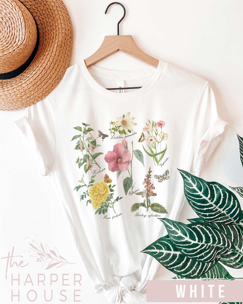 以下が含まれることがあります： 様々な花と蝶の植物画が描かれた白いTシャツ。学名が記載されています。Tシャツはクルーネックで半袖です。「The Harper House」の文字が左下にプリントされています。「WHITE」の文字が右下にプリントされています。