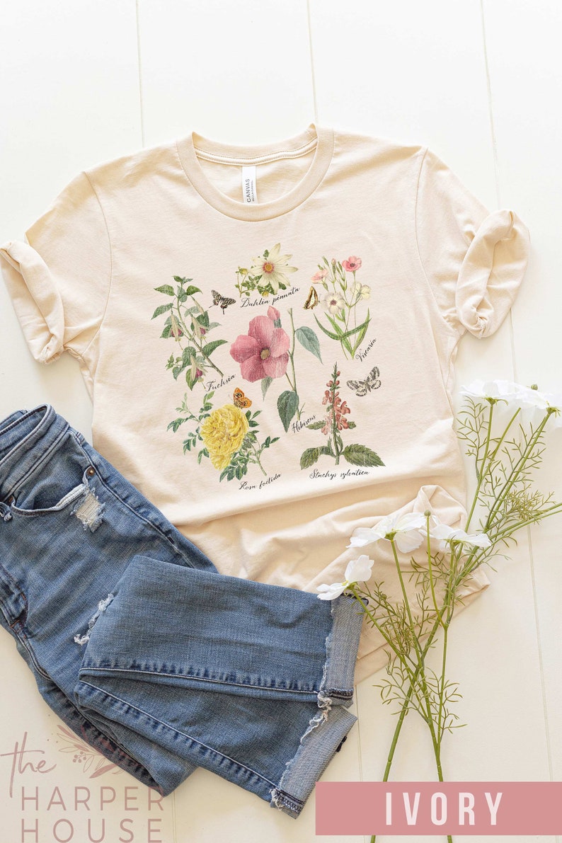 以下が含まれることがあります： 様々な花と蝶の植物画が描かれたアイボリー色のTシャツ。科学名が記載されています。青いデニムジーンズと組み合わせています。The Harper Houseのロゴが見えます。