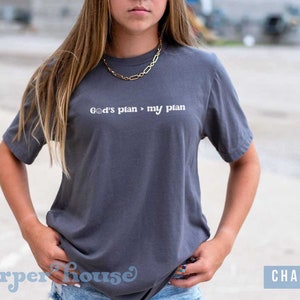 May include: A charcoal gray t-shirt with the text "God's plan > my plan" printed in white.