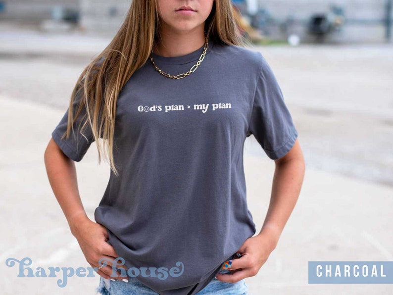 May include: A charcoal gray t-shirt with the text "God's plan > my plan" printed in white.