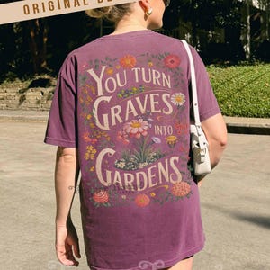 May include: A purple t-shirt with the text "You Turn Graves Into Gardens" in a floral design. The design features various flowers in shades of pink, yellow, and orange. The shirt has short sleeves and a relaxed fit.