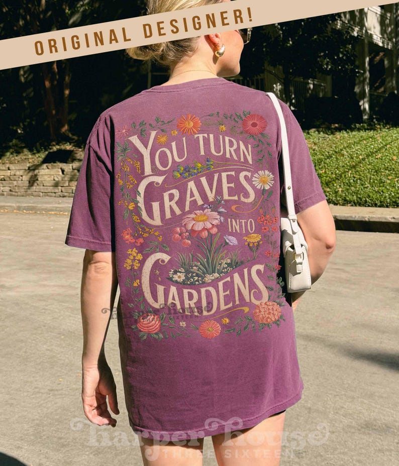 May include: A purple t-shirt with the text "You Turn Graves Into Gardens" in a floral design. The design features various flowers in shades of pink, yellow, and orange. The shirt has short sleeves and a relaxed fit.