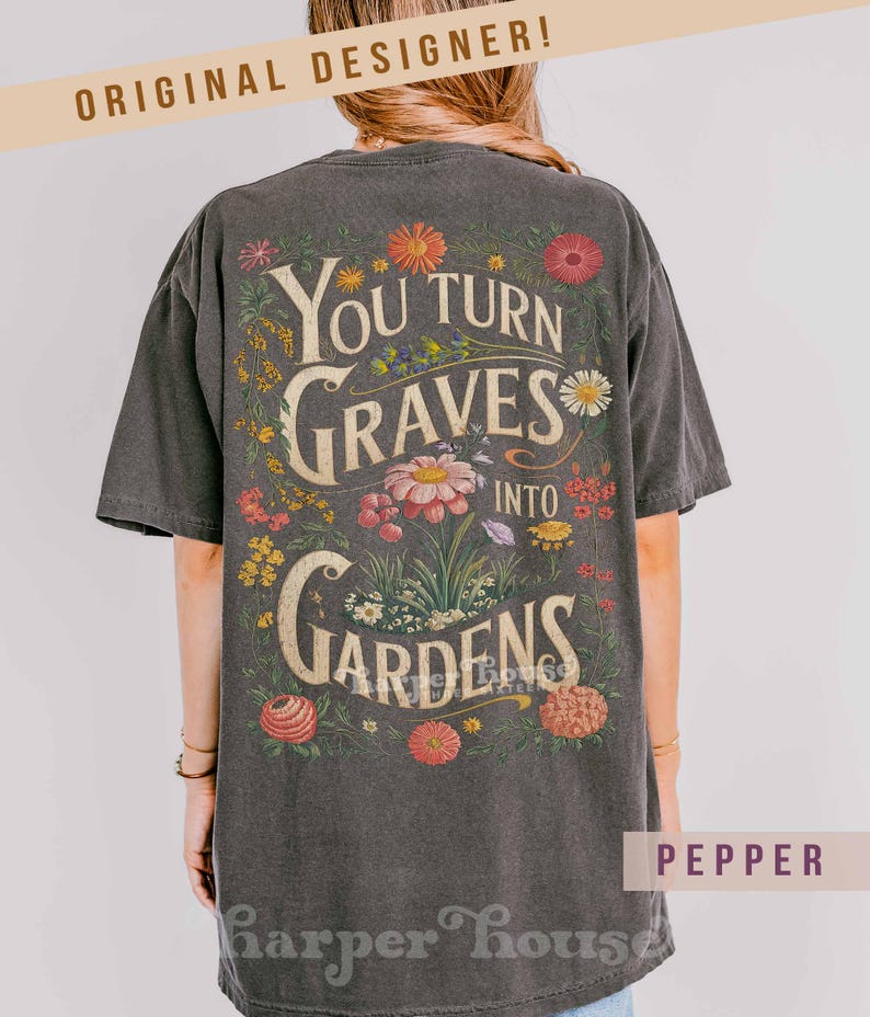 May include: Dark gray t-shirt with the text "You Turn Graves Into Gardens" surrounded by colorful flowers. The shirt has a vintage, worn look and the words "Original Designer" are at the top.